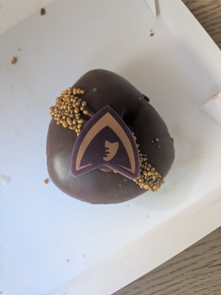 A gordy-as-fuck triangular doughnut with chocolate glaze, gold popping candy sash and traitors logo icing sticker.